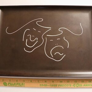 May include: A rectangular, dark brown serving tray with a white line drawing of two theatrical masks, one smiling and one frowning. A wooden ruler is at the bottom.