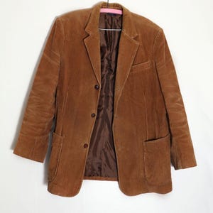 Può includere: Un blazer in velluto a coste marrone con tessuto testurizzato, risvolto dentellato e tre bottoni marrone scuro sul davanti. La giacca presenta due ampie tasche applicate in vita, una tasca sul petto e una fodera interna liscia marrone scuro.