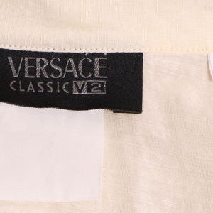 以下が含まれることがあります： クリーム色の生地のクローズアップ。黒いラベルには「VERSACE CLASSIC V2」と書かれています。数字の「40」が書かれた白いタグも表示されています。生地はシャツまたはブラウスのようです。