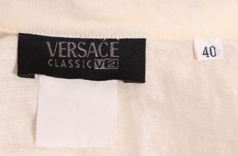 以下が含まれることがあります： クリーム色の生地のクローズアップ。黒いラベルには「VERSACE CLASSIC V2」と書かれています。数字の「40」が書かれた白いタグも表示されています。生地はシャツまたはブラウスのようです。