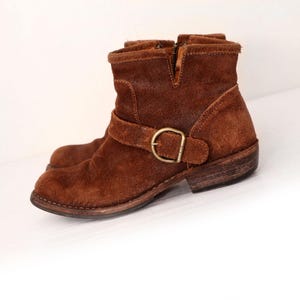 May include: A pair of brown suede ankle boots. The boots have a short shaft, a side zipper, and a strap with a buckle. The soles are a darker brown, and the boots appear worn.