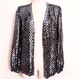 Può includere: Giacca nera con paillettes, maniche lunghe e parte anteriore aperta. La giacca è esposta su un manichino su sfondo bianco. Le paillettes sono argentate e nere, creando un effetto scintillante.