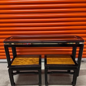 May include: A black lacquered console table with a glass top and gold trim. Two matching black lacquered side tables with wood tops are included.