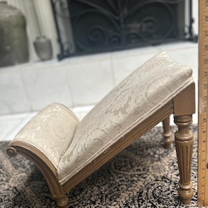 May include: A small, wooden footstool with a beige fabric top. The footstool has a curved back and turned legs.