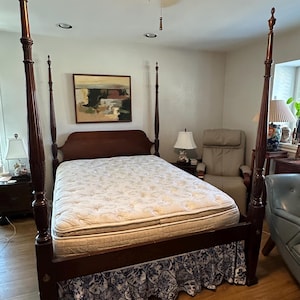 May include: A dark wood four-poster bed with a patterned mattress and a decorative skirt. The bed frame has ornate details and is set in a well-lit room with a painting and a chair.