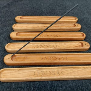 May include: Five wooden incense stick holders with a crescent moon design. The holders are stacked on top of each other and a black incense stick is laying across the top.