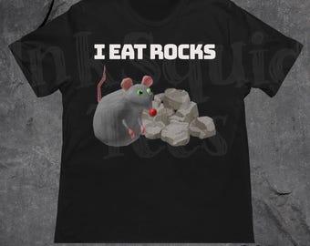 May include: Black t-shirt featuring a cartoon mouse with green eyes and a red nose, next to a pile of rocks. The text "I EAT ROCKS" is printed in white above the mouse and rocks. The shirt has short sleeves.