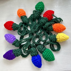 Puede incluir: Una cadena de bombillas de luz de ganchillo en varios colores, incluyendo rojo, naranja, amarillo, verde, azul y morado. Las bombillas están conectadas por hilo verde.