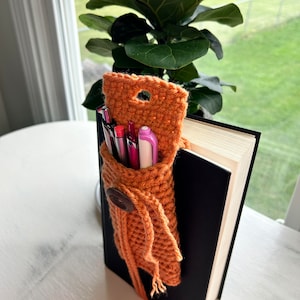 May include: A crocheted orange book holder with a brown button and a tassel. The holder is filled with pens and pencils and is attached to a black book.