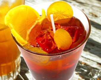 May include: A vibrant cocktail in a clear glass, garnished with orange slices and a small orange fruit. The drink has a deep red hue, with ice cubes and a white straw. The words "CUTIE ART CANDLE" are visible in the background.