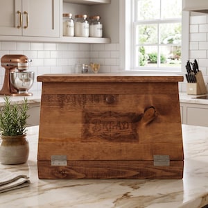 May include: A medium-brown wooden bread box with a slanted front panel featuring the word BREAD engraved in a decorative frame. The box has a flat top, a hinged front door with a small round knob, and two visible silver-toned metal hinges.