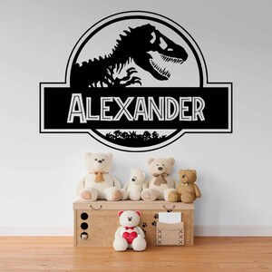 May include: Black and white dinosaur skeleton decal with the name "ALEXANDER" in a black banner below. The decal is on a white wall with a wooden chest and teddy bears in front of it.