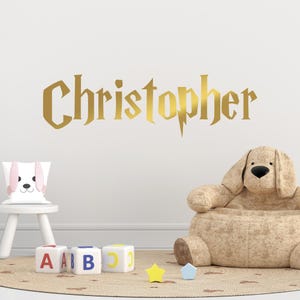 May include: Gold glitter vinyl wall decal with the name "Christopher" in a Harry Potter-inspired font. The decal is displayed on a white wall above a child's play area with a stuffed animal chair, alphabet blocks, and a star-shaped rug.