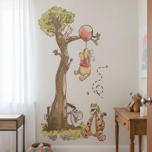 May include: A whimsical wall decal featuring Winnie the Pooh and friends. The illustration shows Pooh hanging from a red balloon, Piglet in a tree, Tigger and Eeyore. The artwork is in a soft, watercolor style, perfect for a nursery or child's room.