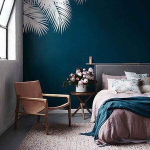 Könnte beinhalten: Ein blaugrünes Schlafzimmer mit einem Bett, einem Stuhl, einem Beistelltisch und einem Teppich. Die Wand hat ein weißes Palmenbaum-Dekor. Das Bett hat eine rosa Decke und eine blaugrüne Überwurfdecke. Der Stuhl ist aus Leder und Holz. Der Beistelltisch ist aus Holz und hat eine Vase mit rosa Blumen darauf. Der Teppich ist aus hellbraunem Material.