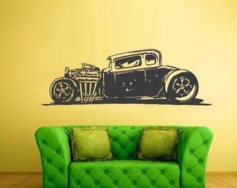 May include: A dark grey silhouette of a classic hot rod car is displayed on a yellow wall. The car features large wheels, an exposed engine, and a vintage body style. Below the car is a green tufted sofa with two green square cushions.