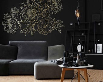 May include: A gold-colored floral wall decal featuring a peony design. The decal is applied to a black wall, with a gray sofa and a small table with decorative items in the foreground. The room has a modern, minimalist aesthetic.