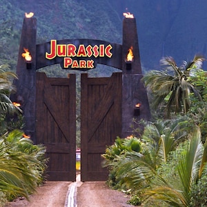 May include: The iconic entrance to Jurassic Park, featuring large wooden gates and a sign with the film's logo. The scene is set in a lush, tropical environment with palm trees and dense foliage, with flames atop the gate pillars.