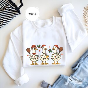 May include: White sweatshirt with a Christmas design of three geese wearing Santa hats and fairy lights. Each goose holds a wooden spoon or a gingerbread cookie. The word "WHITE" is in a white circle.