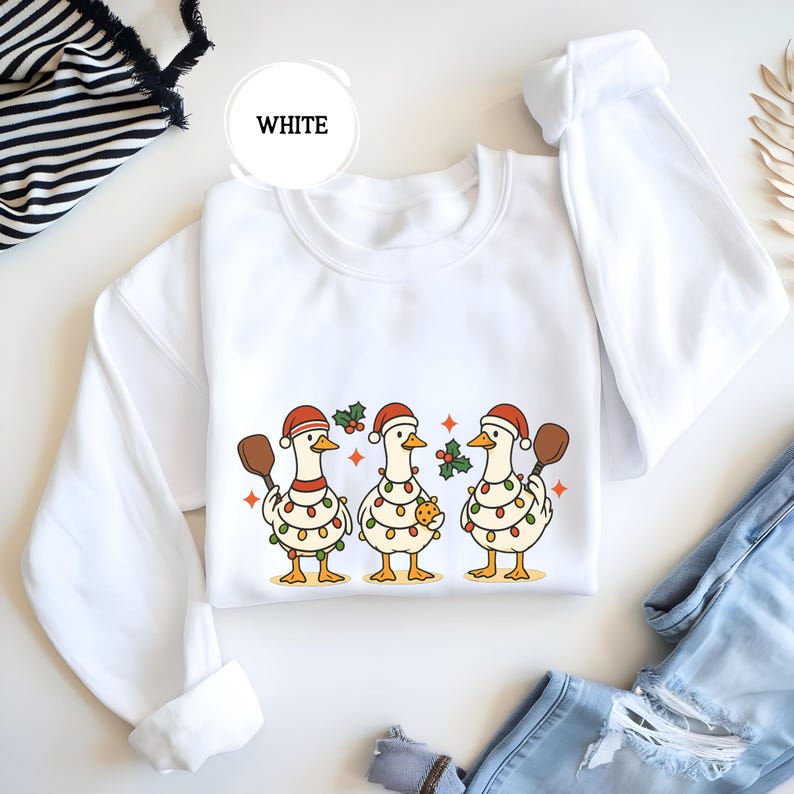 May include: White sweatshirt with a Christmas design of three geese wearing Santa hats and fairy lights. Each goose holds a wooden spoon or a gingerbread cookie. The word "WHITE" is in a white circle.