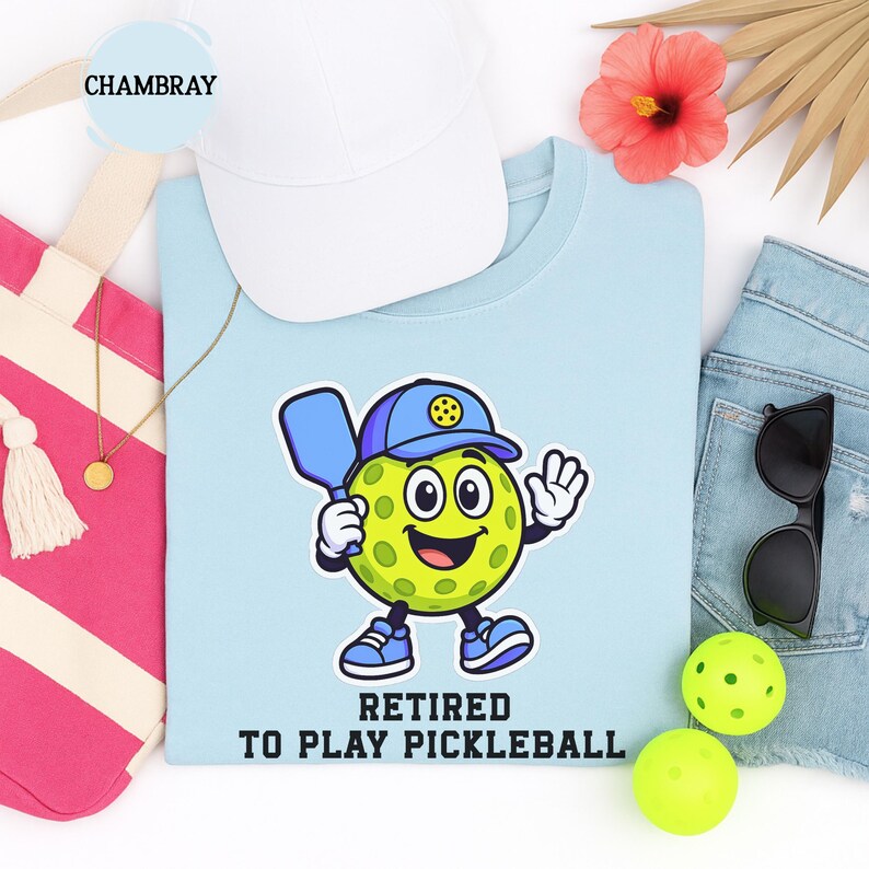 May include: Light blue t-shirt with a cartoon pickleball character holding a paddle and wearing a cap. The text "Retired to Play Pickleball" is printed below the character. A white cap, pink tote bag, and sunglasses are also visible.