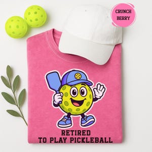 May include: A pink sweatshirt with a cartoon pickleball character wearing a blue cap and holding a paddle. The text "Retired to Play Pickleball" is below the character. A white cap and two green pickleballs are also in the image.