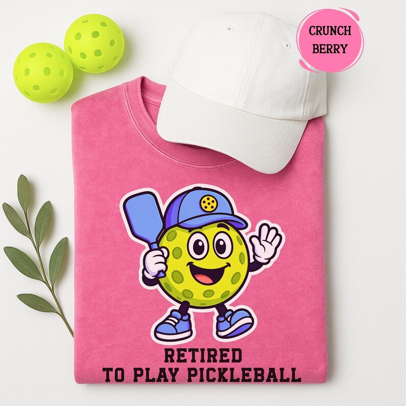 May include: A pink sweatshirt with a cartoon pickleball character wearing a blue cap and holding a paddle. The text "Retired to Play Pickleball" is below the character. A white cap and two green pickleballs are also in the image.