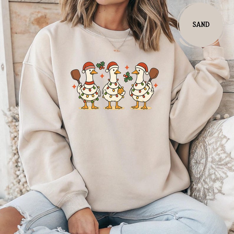 May include: Sand-coloured sweatshirt with a festive design of three cartoon geese wearing Santa hats and string lights. Each goose holds a wooden spoon and some have holly or a cookie. The word "SAND" is in a circle in the upper right.
