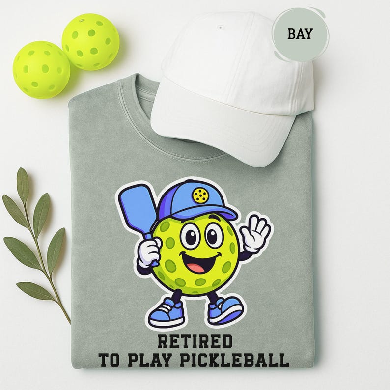 May include: A light green sweatshirt with a cartoon pickleball character wearing a blue cap and holding a paddle. The text "RETIRED TO PLAY PICKLEBALL" is below the character. A white baseball cap and two yellow pickleballs are also in the image.