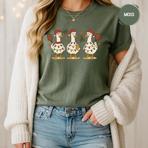 May include: A moss green t-shirt with a festive design of three geese in Santa hats, each holding a paddle and decorated with Christmas lights. One goose holds a biscuit. The shirt is worn by a person, with the word 'MOSS' visible.