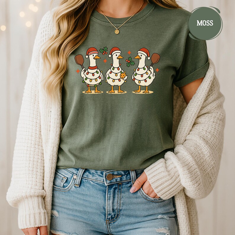 May include: A moss green t-shirt with a festive design of three geese in Santa hats, each holding a paddle and decorated with Christmas lights. One goose holds a biscuit. The shirt is worn by a person, with the word 'MOSS' visible.
