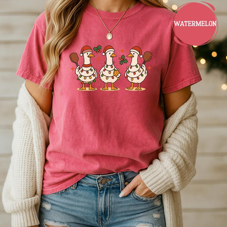 May include: A pink t-shirt with a playful design of three geese adorned with Santa hats and fairy lights. One goose holds a cookie, another a wooden spoon. The word "WATERMELON" is in a pink circle.