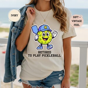 May include: Ivory-colored t-shirt featuring a cartoon pickleball character wearing a blue hat and shoes, holding a paddle. The text "RETIRED TO PLAY PICKLEBALL" is printed below the character. The shirt has a soft, vintage feel.