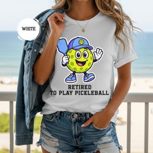 May include: White t-shirt with a cartoon pickleball character wearing a blue hat and holding a paddle. The text "Retired to Play Pickleball" is printed below the character. The shirt is short-sleeved.