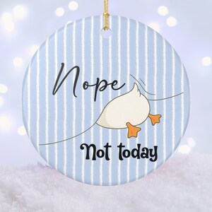 Könnte beinhalten: Runder Keramikschmuck mit blau-weiß gestreiftem Hintergrund. Der Schmuck zeigt eine Cartoon-Ente mit orangefarbenen Füßen und die Schriftzüge "Nope" und "Not today" in schwarzer Schrift. Ein goldener Faden ist zum Aufhängen befestigt.