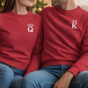 May include: Two red sweatshirts with white crown and initial designs. One sweatshirt has a "Q" and the other has a "K". The sweatshirts are paired with blue jeans. The image shows a couple wearing the matching sweatshirts.