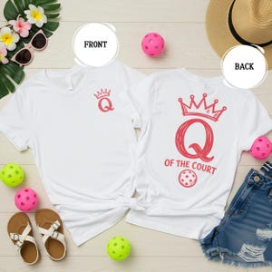 May include: White t-shirts with a red crown and the letter "Q" design. One shirt has "Q" with a crown on the front. The other shirt has "Q OF THE COURT" with a pickleball graphic on the back. Accessories include sunglasses, sandals, and pickleballs.