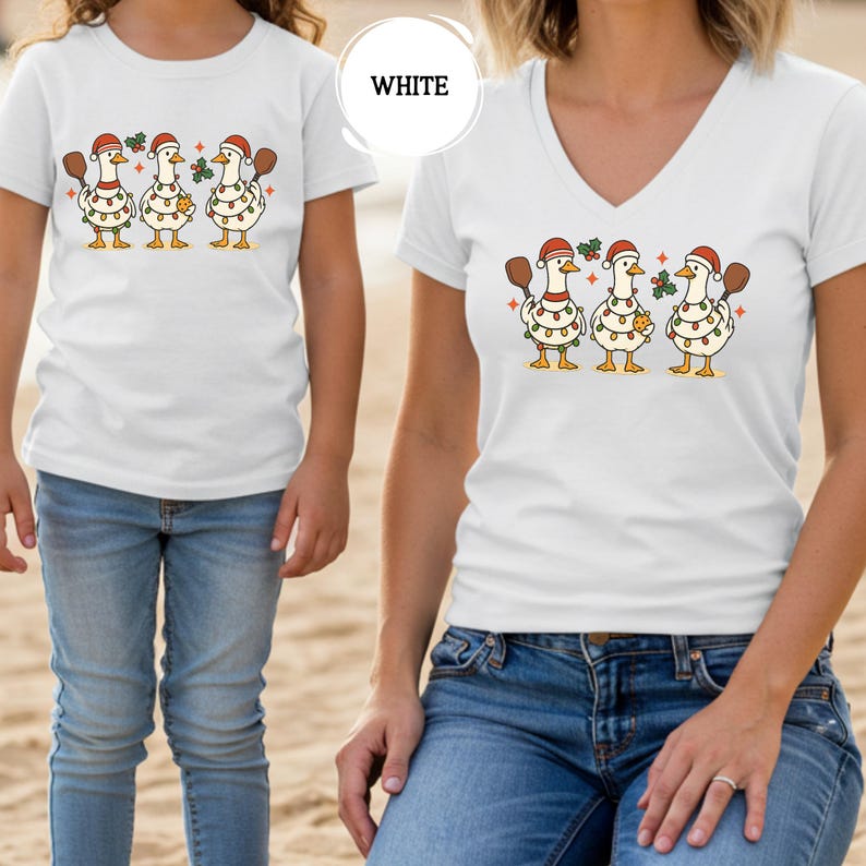 May include: White t-shirts featuring a graphic of three geese wearing Santa hats and Christmas lights, holding paddles. The word "WHITE" is in a white circle. The shirts are worn by a child and an adult, both wearing blue jeans.