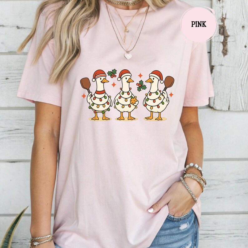 May include: Pale pink t-shirt with a festive design of three geese in Santa hats, each holding a paddle. They are decorated with fairy lights and holly. The illustration has a retro cartoon style.