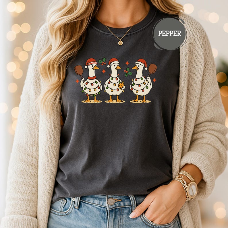 May include: Dark grey t-shirt with a festive design of three geese wearing Santa hats and fairy lights. Each goose holds an item: a spatula, a cookie, and a football. The word "PEPPER" is printed on the shirt.