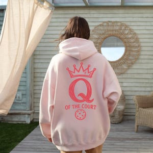 May include: A light pink hoodie with a hood, featuring a coral-colored graphic on the back. The graphic includes a crown, the letter "Q", the words "OF THE COURT", and a pickleball. The hoodie is made of a soft material.