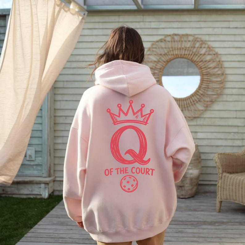 May include: A light pink hoodie with a hood, featuring a coral-colored graphic on the back. The graphic includes a crown, the letter "Q", the words "OF THE COURT", and a pickleball. The hoodie is made of a soft material.