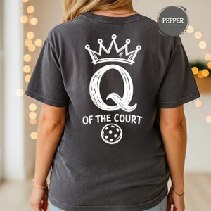 May include: Dark gray t-shirt with a white graphic on the back. The graphic includes a crown, a large letter "Q", the words "OF THE COURT", and a pickleball. The shirt is labeled "Pepper".