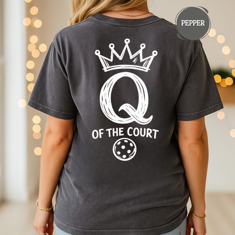 May include: Dark gray t-shirt with a white graphic on the back. The graphic includes a crown, a large letter "Q", the words "OF THE COURT", and a pickleball. The shirt is labeled "Pepper".