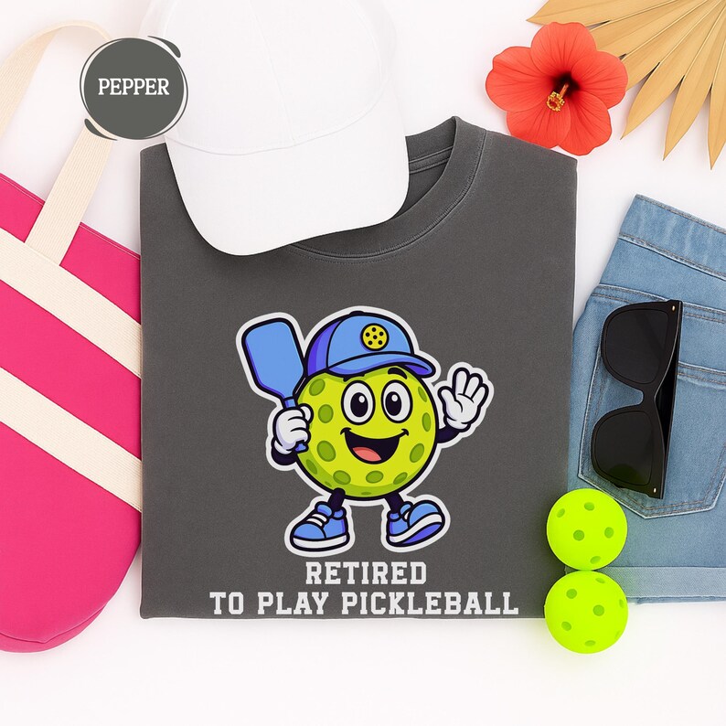 May include: A charcoal gray t-shirt featuring a cartoon pickleball character with a blue hat and paddle, with the text "RETIRED TO PLAY PICKLEBALL". The image also includes a white cap, sunglasses, and two pickleballs.