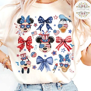 Peut inclure: Un t-shirt blanc &agrave; th&egrave;me patriotique rouge, blanc et bleu avec Mickey Mouse, Minnie Mouse, Donald Duck et Dingo portant des lunettes de soleil et des chapeaux rouge, blanc et bleu. Le t-shirt pr&eacute;sente &eacute;galement des n&oelig;uds et des &eacute;toiles rouge, blanc et bleu.