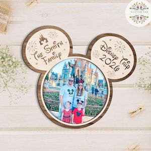 May include: A wooden Mickey Mouse-shaped photo frame with a family photo inside. The frame has the text "The Smith Family" and "Disney Trip 2026" on the ears. The frame is on a light wood surface.