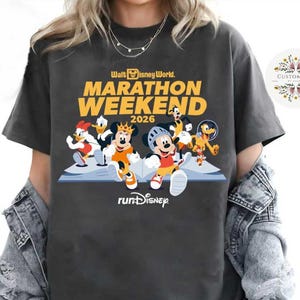 Peut inclure: T-shirt gris foncé avec un motif de personnages Disney en train de courir. Le t-shirt affiche le texte "Walt Disney World Marathon Weekend 2026" en jaune et blanc. Les mots "runDisney" sont également imprimés sur le t-shirt.