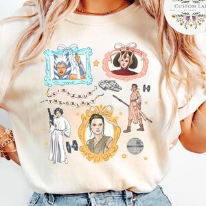 May include: Cream-colored t-shirt featuring framed illustrations of female characters from the Star Wars franchise. The phrase "Girls Run The Galaxy" is written in a stylized font. The shirt has a round neck and short sleeves.