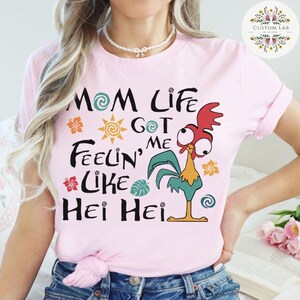 May include: Pink t-shirt with a cartoon rooster and the text "Mom Life Got Me Feelin' Like Hei Hei".
